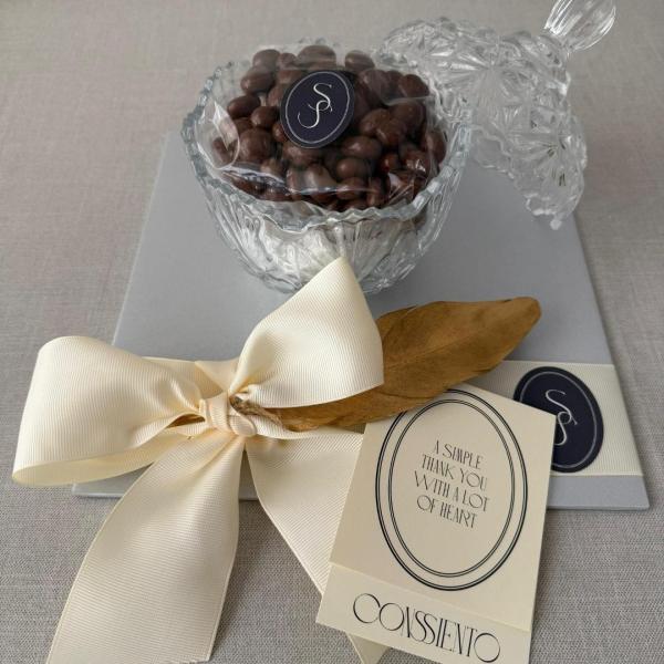 BOWL CON BOTANA DULCE O SALADA, con tapa transparente de CONSIIENTO, relleno de pasas cubiertas de chocolate, presentado con moño en tono marfil y tarjeta personalizada, ideal como regalo elegante.