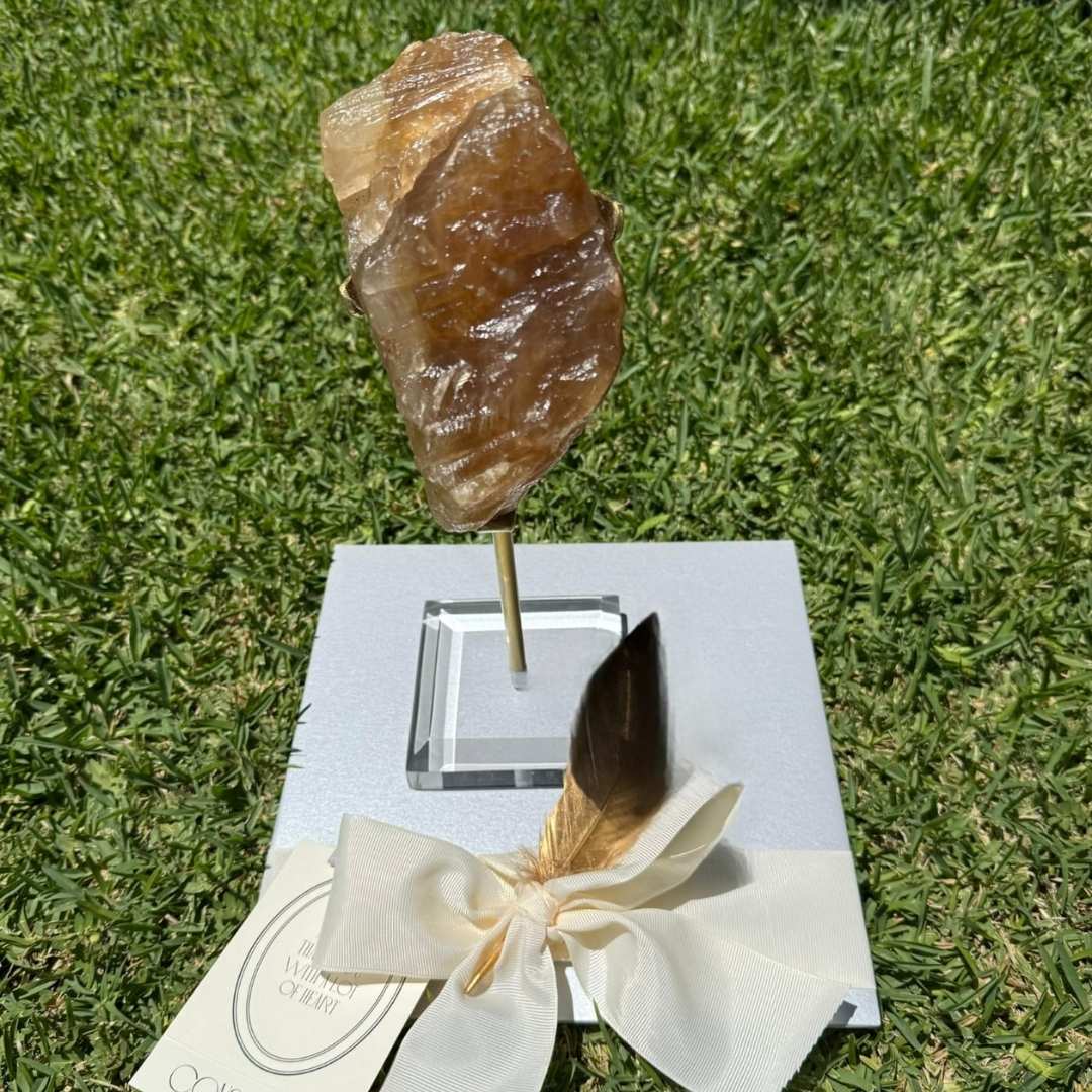 CALCITA CON PEDESTAL en tonos marrón ámbar montada en pedestal dorado con base acrílica transparente, creación de SSIENTO que resalta su forma irregular como pieza decorativa elegante y sofisticada. Esta pieza puede variar en forma, tamaño y tonos.